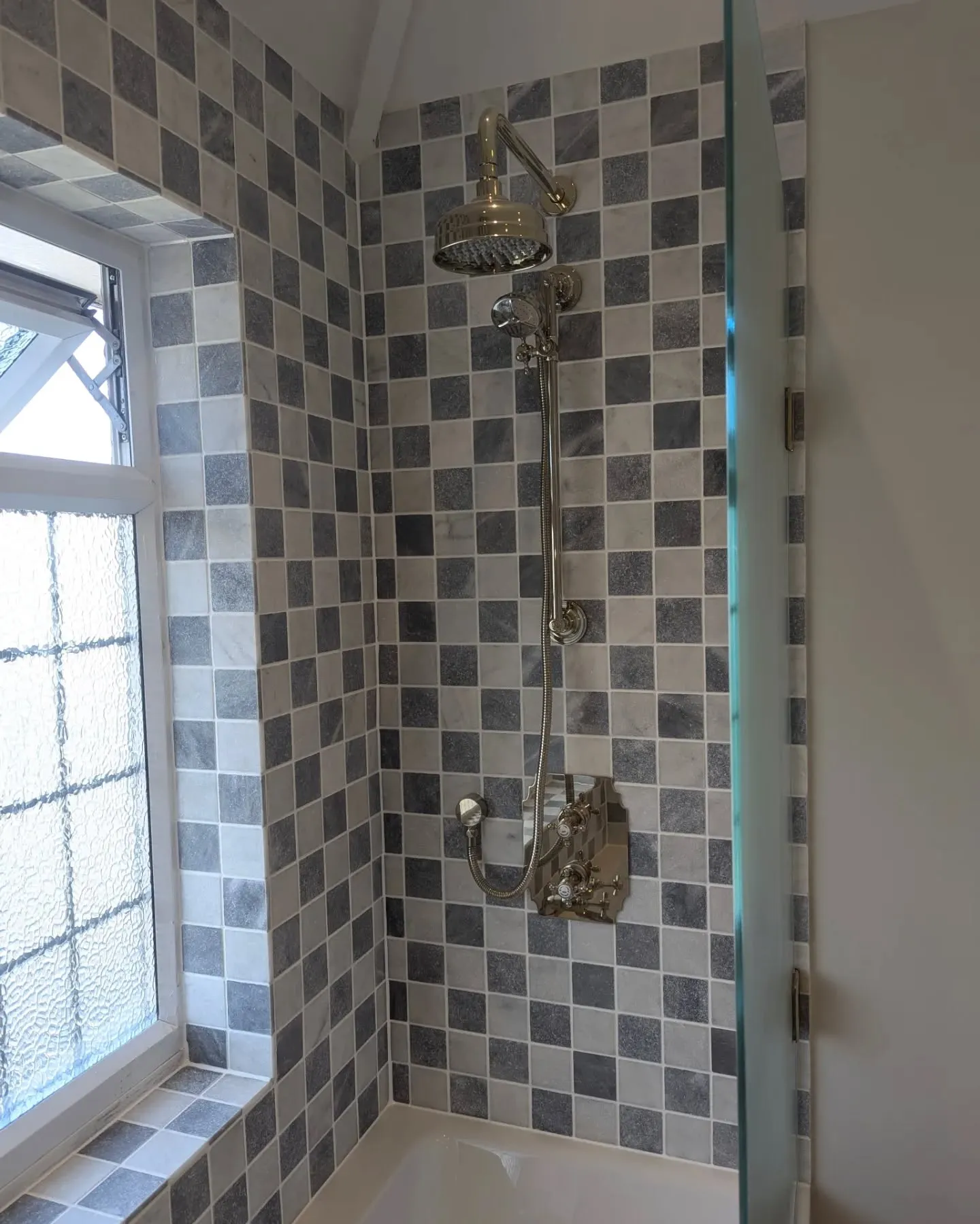 A bath and shower with chequered tiles and a window.